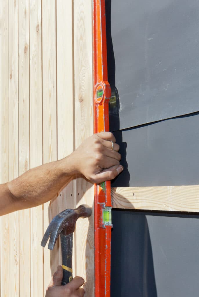 Pose Bardage Bois : Utilisation Niveau à Bulle Un ouvrier ajuste un niveau à bulle orange contre un pare-pluie noir lors de la pose d'un bardage en bois clair vertical.