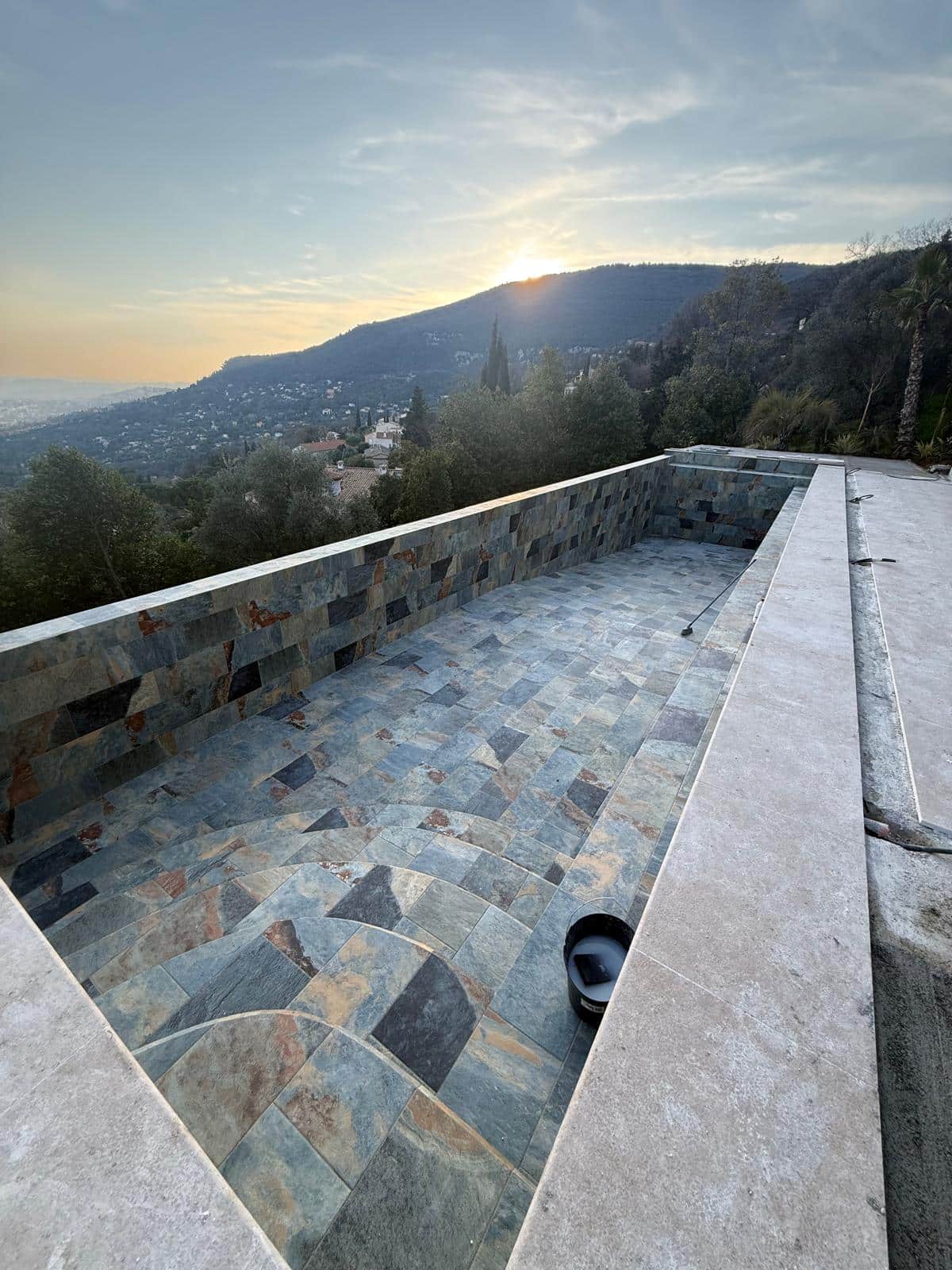 Piscine en Ardoise avec Vue Panoramique sur la Montagne Piscine en construction avec carrelage en ardoise multicolore et vue panoramique sur les montagnes au coucher du soleil.